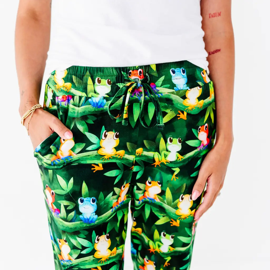 Close-up of a woman wearing dark green pajama pants with a colorful tree frog and jungle leaf print and a drawstring waist.